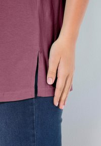 Maglietta marrone con spacchi laterali, maniche corte e una consistenza morbida. Abbinata a jeans in denim scuro. Mano posizionata vicino all'orlo.
