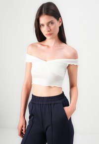Hvit av-skulder crop top med volanger i kantene, med et vridd design. Kombinert med marineblå bukser med elastisk midje, hender i lommene.