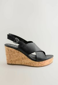 Black wedge sandal with crisscross leather straps, cork-textured heel, and adjustable ankle strap. Smooth footbed and flat rubber sole.