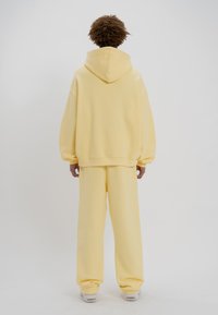 Sudadera amarilla de gran tamaño y pantalones a juego fabricados en material suave, con una gran capucha y corte relajado, combinados con zapatillas de color claro.