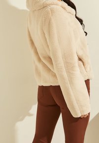 Beżowy kaptur z futra syntetycznego o przytulnej fakturze, zestawiony z dopasowanymi brązowymi spodniami. Projekt charakteryzuje się luźnym krojem i miękkim materiałem.