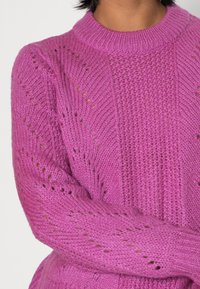 Stickad tröja i klara rosa med ribbat rundhals, öppna stickmönster och långa ärmar. Mjuk textur med detaljer på kroppen och ärmarna.