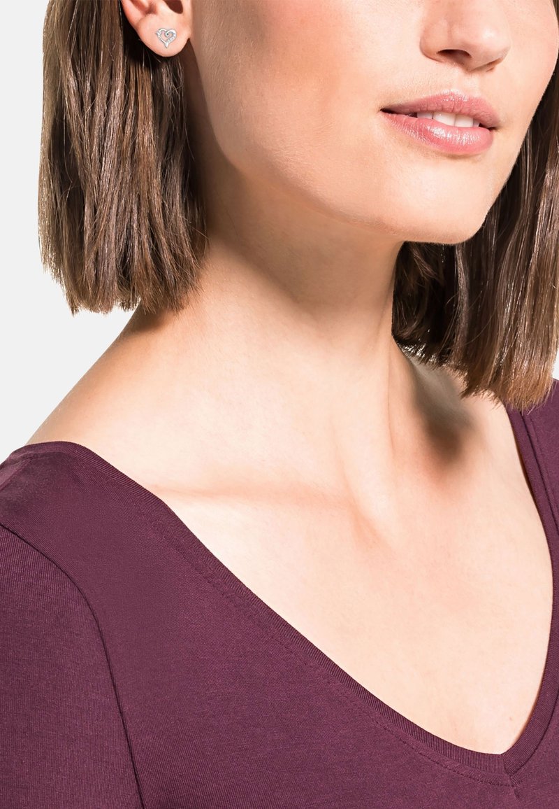 Silverfärgade hjärtformade studs som bärs av en modell med rakt brunt hår, tillsammans med en djup burgundy v-ringad topp. Slät textur synlig.