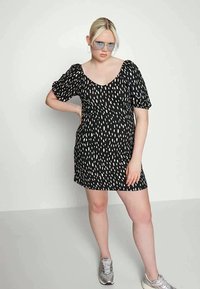 Vestido preto com padrão abstrato branco, mangas curtas com puff, decote em forma de scoop, comprimento até ao joelho, tecido leve, combinado com ténis prateados.
