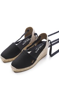 Sandali con zeppa nera in tela con suola in juta intrecciata, dotati di cinghie alla caviglia e una texture liscia. Logo dorato in rilievo sulla soletta.