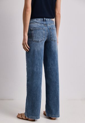 Personne portant un jean bleu taille haute à jambes larges et un top noir sans manches, debout pieds nus devant un fond clair uni.