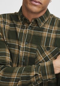 Camisa de franela a cuadros verdes y marrones con cuello puntiagudo, botones frontales y un bolsillo en el pecho, con una textura suave y líneas blancas contrastantes.