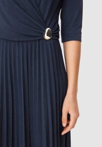 Abito plissettato blu navy con una texture liscia, caratterizzato da un corpetto attorcigliato e un dettaglio di accessorio dorato in vita.