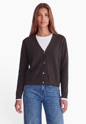 Femme portant un cardigan boutonné marron foncé sur une chemise blanche et un jean bleu, debout devant un fond clair uni.
