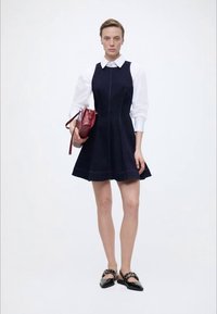 Γυναίκα που φοράει μπλε σκούρο αμάνικο φόρεμα πάνω από λευκό πουκάμισο με γιακά, κρατώντας μπορντό τσάντα και φορώντας μαύρα επίπεδα παπούτσια με λουράκια.