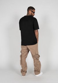 Čierne bavlnené tričko a khaki cargo nohavice s viacerými vreckami, doplnené o biele tenisky. Pozadie je jednoduchá svetlá farba.