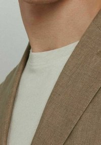 Blazer marrone chiaro con tessuto strutturato, abbinato a una T-shirt grigia chiara tinta unita. Il blazer ha ampi revers e cuciture visibili.