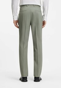 Homme portant un pantalon de costume gris clair et des chaussures de ville noires, debout de dos devant un fond blanc.