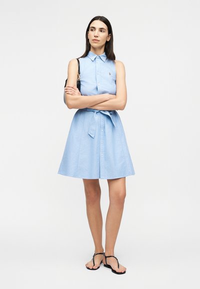 Sieviete ar gariem tumšiem matiem, tērpusies gaiši zilā bezroku pogātā kleitā ar jostu viduklī, melnās sandales un melnu plecu somu, stāvot ar sakrustotām rokām.