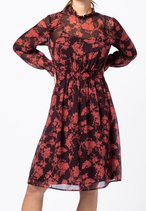 Femme portant une robe noire transparente avec un motif floral rouge, manches longues, col volanté et taille cintrée, debout les mains sur les hanches.