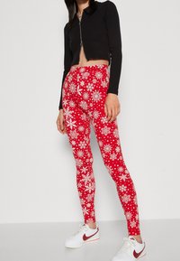 Rode leggings met een wit sneeuwvlokpatroon, gemaakt van rekbare stof. Gecombineerd met een zwarte crop-top met ritssluiting en witte sneakers.