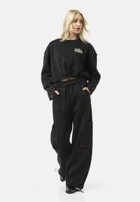 Zwarte cropped sweatshirt en brede sweatpants, beide met elastische details. De broek heeft grote klepzakken met rode accenten. Gladde textuur.