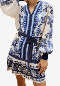 Vestito stampato floreale in bianco e blu con scollatura a V, lunghe maniche a palloncino, vita cintrata e motivi intricati lungo l'orlo.