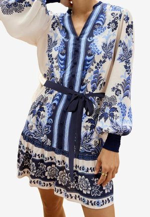 Bloemenprint jurk in wit en blauw met een V-hals, lange puffmouwen, een ceintuur in de taille en ingewikkelde patronen langs de zoom.