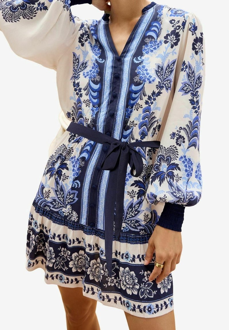 Vestito stampato floreale in bianco e blu con scollatura a V, lunghe maniche a palloncino, vita cintrata e motivi intricati lungo l'orlo.