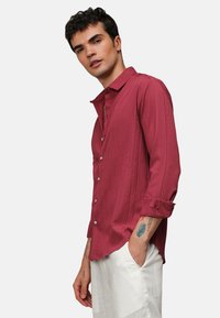 Camicia lunga rossa con texture e bottoni, caratterizzata da un colletto rilassato e maniche arrotolate, indossata sopra pantaloni bianchi.
