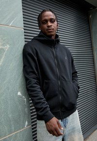 Sort zip-up hoodie lavet af blødt stof, med en frontlomme og et lille Jordan-logo på brystet. Båret med lyseblå denimjeans.