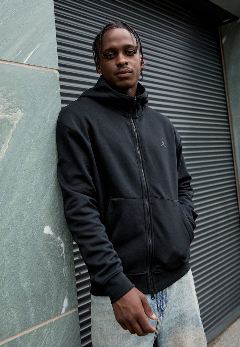 Sort zip-up hoodie lavet af blødt stof, med en frontlomme og et lille Jordan-logo på brystet. Båret med lyseblå denimjeans.