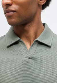 Κοντινό πλάνο ενός ατόμου που φοράει ένα ανοιχτοπράσινο πόλο με υφή και γιακά σε σχήμα V, κοιτώντας ελαφρώς προς τα πλάγια.