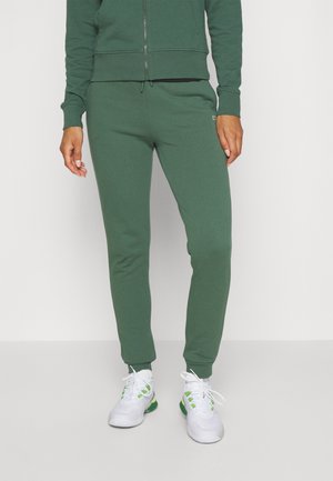 Personne portant un pantalon de jogging vert et une veste zippée assortie avec des chaussures de sport blanches et vertes, debout sur un fond uni.