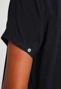 Sort kortærmet skjorte med en blød tekstur, med en rullet ærme fastgjort af en hvid knapdetalje. Minimalistisk design og stof, der hænger.