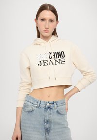Členková béžová mikina s kapucňou na šnúrku a grafickým textom "Moschino Jeans". V kombinácii s svetlomodrými džínsy s vysokým pásom.