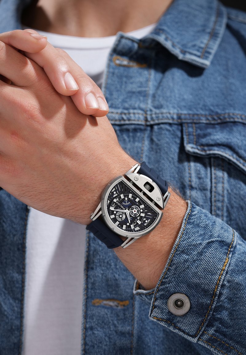 Moški nosi modro silikonsko zapestnico s črnim in srebrnim geometrijskim številčnico ter delno pokrito krono na levi roki.