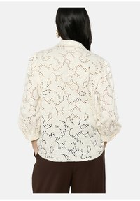 Blusa bianca in broderie anglaise con maniche a sbuffo, motivo intagliato intricato e orlo rotondo, caratterizzata da una texture leggera.