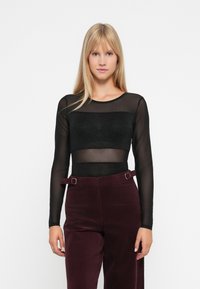 Una donna con lunghi capelli biondi indossa un top trasparente nero a maniche lunghe con un bandeau nero e pantaloni a vita alta di un viola scuro con dettagli in cintura.