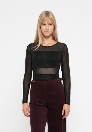 Una donna con lunghi capelli biondi indossa un top trasparente nero a maniche lunghe con un bandeau nero e pantaloni a vita alta di un viola scuro con dettagli in cintura.