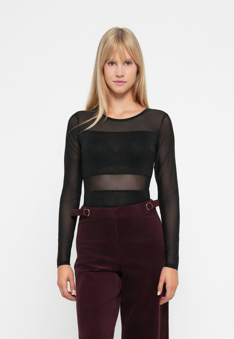 Una donna con lunghi capelli biondi indossa un top trasparente nero a maniche lunghe con un bandeau nero e pantaloni a vita alta di un viola scuro con dettagli in cintura.