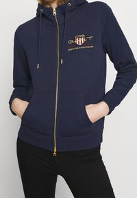 Námořnická modrá mikina na zip z bavlněné směsi. Obsahuje přední logo, zlatý zip a dvě přední kapsy. Žebrované manžety a spodní lem.