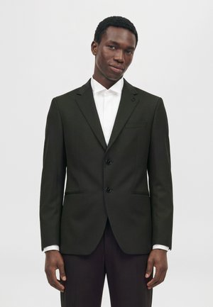 Un homme porte un costume noir à deux boutons et une chemise blanche sur un fond clair et uni.