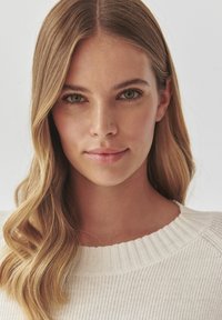 Portræt af en person med langt, bølget blonde hår, iført en lys, ribbet sweater med en struktureret hvid halsudskæring, sat mod en neutral baggrund.