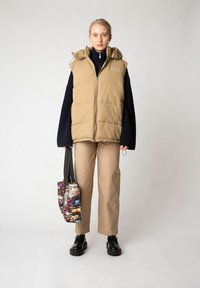 Khaki puffer-väst med dragkedja och huva, bärs över en marinblå tröja, matchad med beige byxor och en flerfärgad mönstrad tygväska.