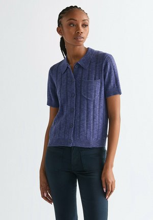 Krótki sweter na guziki z krótkim rękawem wykonany z teksturowanego fioletowego dzianiny, z kieszenią i pionowym prążkowanym wzorem, w połączeniu z ciemnymi spodniami.