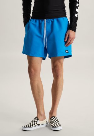 Mand iført klare blå shorts med snøre, sort langærmet trøje og sort-hvide ternede slip-on sko, stående med hænderne ned langs siderne.