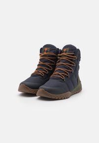 Bottes de randonnée navy avec lacets marron, semelle en caoutchouc et tige en toile. Présente un talon texturé vert et un patch logo sur la languette.
