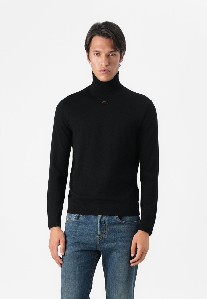 Μαύρο πουλόβερ ζιβάγκο με μακριά μανίκια, εφαρμοστή σχεδίαση και μικρή λεπτομέρεια λογότυπου στον λαιμό, συνδυασμένο με μπλε τζιν.