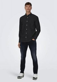 Jovem em pé usando uma camisa preta abotoada, jeans azul escuro e sapatilhas brancas contra um fundo claro e simples.