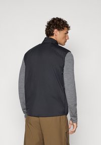 Zwarte mouwloze vest met hoge kraag, van lichtgewicht stof en met een gladde textuur, gedragen over een grijs langemouwenshirt.