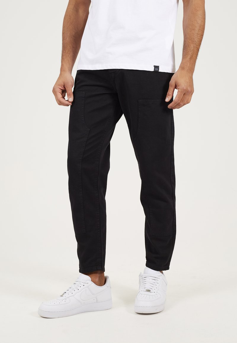 Pantalones negros ajustados con una textura suave, que cuentan con bolsillos frontales y una cinturilla ligeramente elástica, combinados con zapatillas blancas.