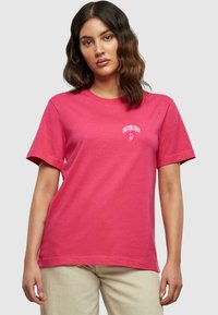 Rosa bomull T-skjorte med korte ermer og rund hals, med en liten hvit grafisk tekstdesign på øvre venstre side.