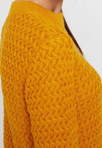 Gul stickad tröja med ett texturerat mönster och ribbad krage, som visar en närbild av ärmen och axelområdet.