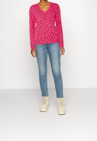 Maglione rosa a scollo a V con pois neri, abbinato a jeans blu e stivaletti beige con lacci. Il maglione presenta maniche lunghe e una vestibilità ampia.